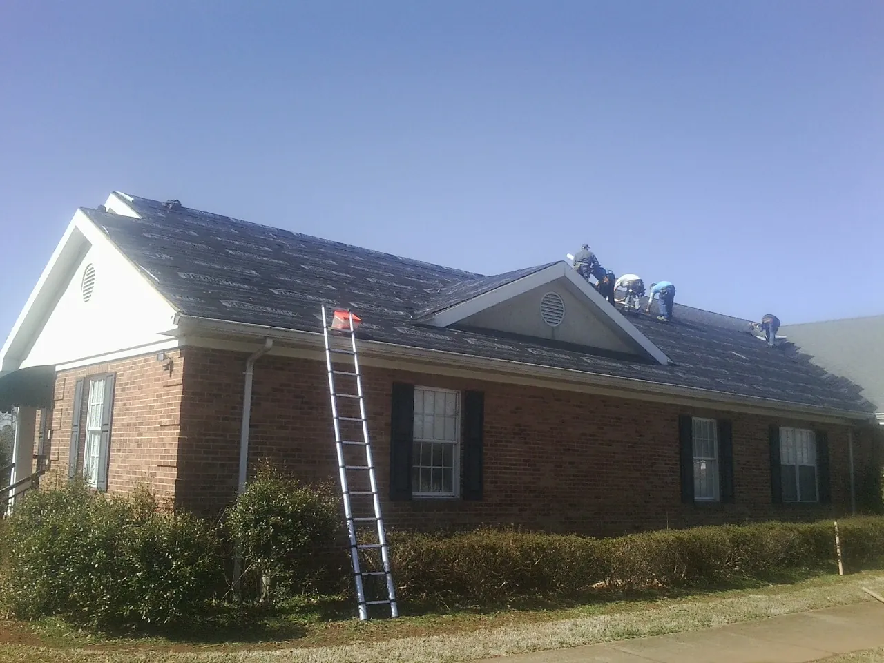 Crew tearing shingles off a brick ranch roof with a ladder set — Emergency Roof Repair work by our crew in North Greece