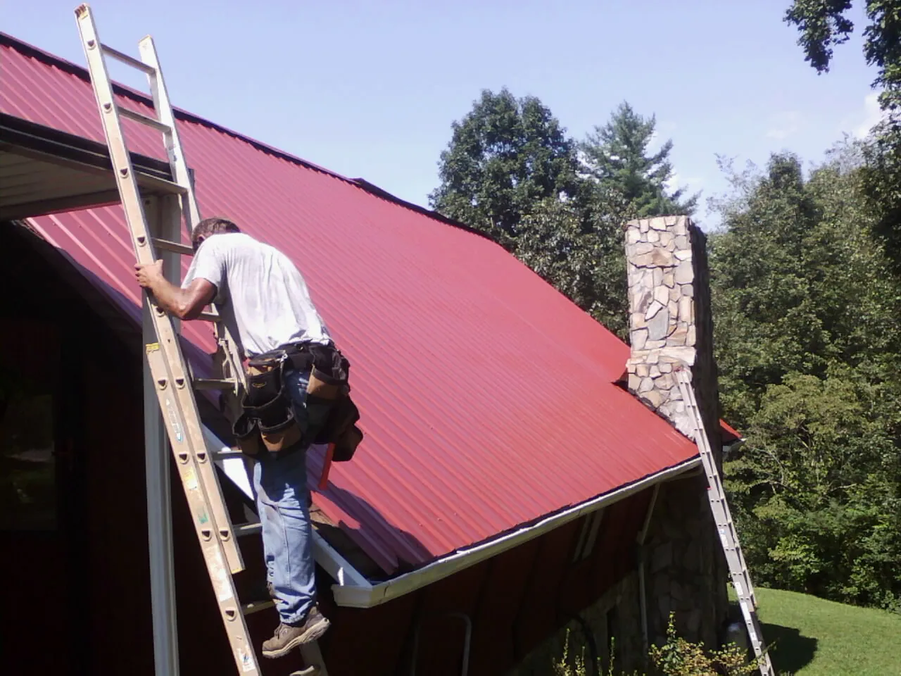 Roofer on a ladder beside a red metal roof and stone chimney — Soffit & Fascia Repair by Roofing Brothers in North Greece