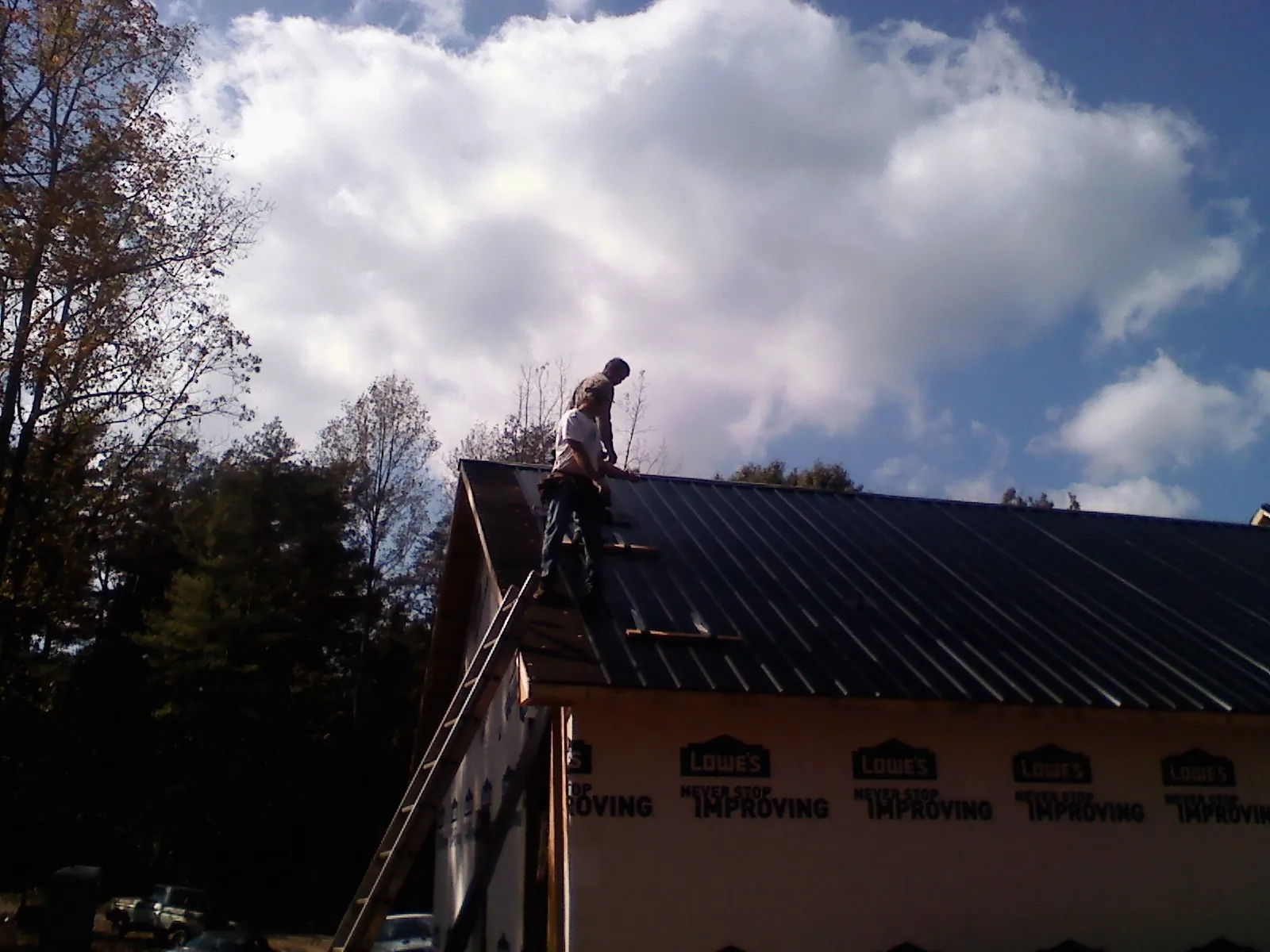 Roofer fastening dark metal panels high against a cloudy sky, from a Roof Inspection job in North Greece